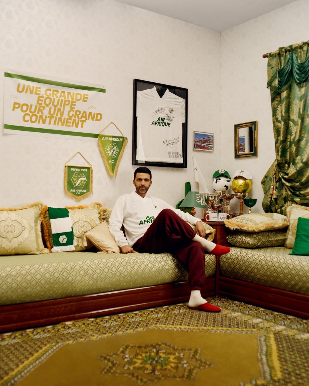 Hicham El Guerrouj, vêtu d’un élégant sweat Air Afrique, assis fièrement dans son salon marocain au décor traditionnel, regard confiant et posture sereine.